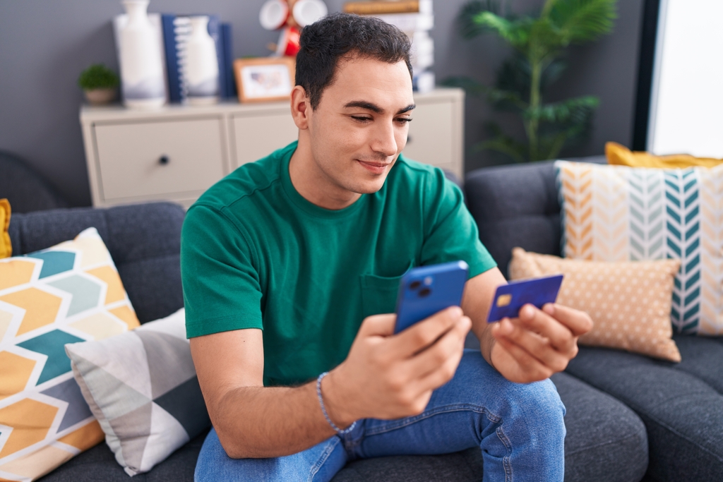 Joven hispano usando smartphone y tarjeta de crédito sentado en un sofá en casa